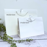 Two plain white paper gift bags with white handles, shown on a light rustic surface with green eucalyptus stems. Black text labels indicate dimensions: 44x32x12cm on the larger bag and 31x22x12cm on the smaller bag.