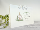 A white paper gift bag with white handles, printed with a watercolor church illustration and the personalized text 'ON YOUR CHRISTENING DAY Lucas David Smith 23/08/206'. The bag is photographed on a light gray wood surface with green trailing leaves at the left edge.